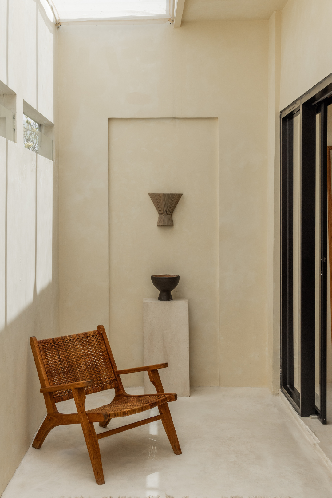 Wooden armchair in a modern interior setting with light-colored walls and a large window and wall light in clay on the wall.