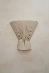 A bone-colored cloth wall light sconce designed to be mounted on a wall, with a textured surface and a conical shape at the bottom.