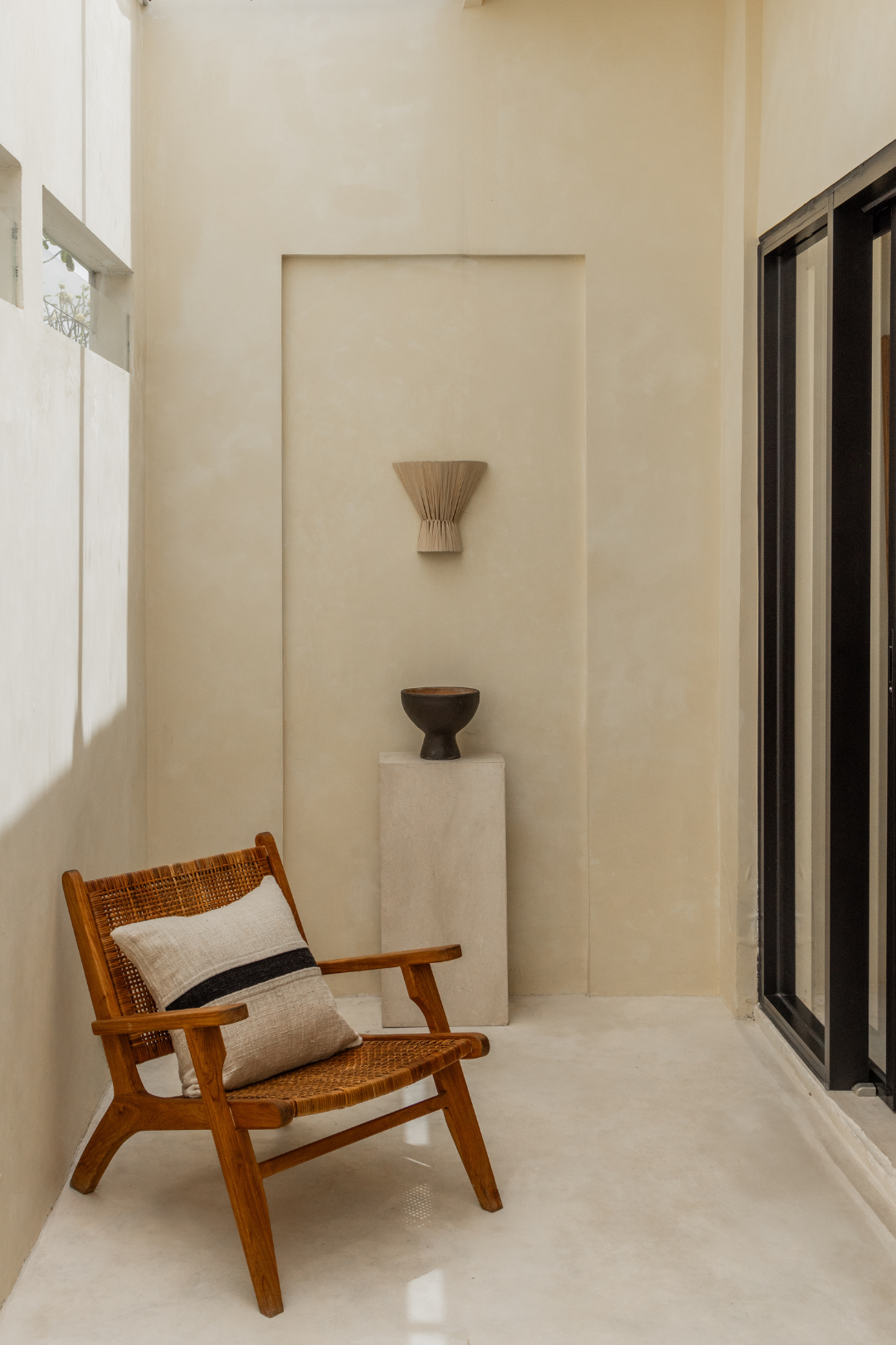 Wooden armchair with a pillow against a beige wall with decorative vases and flume wall light hanging on the wall.