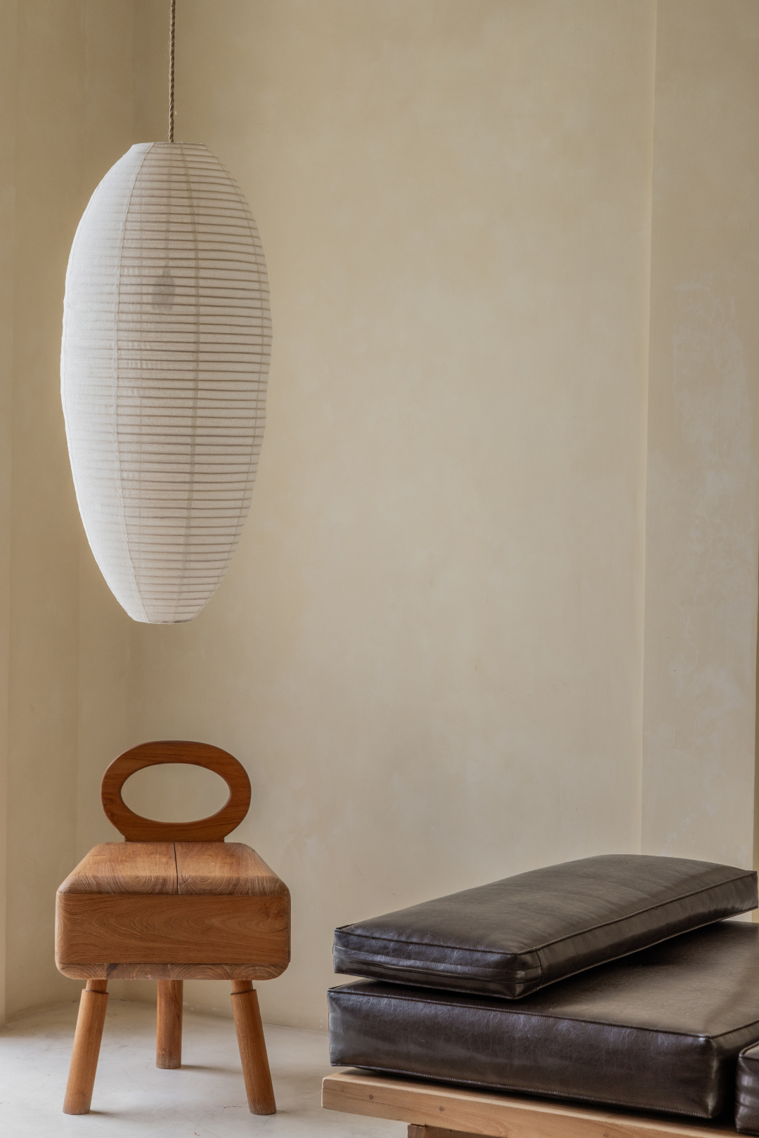 White pendant lamp hanging above a wooden stool and cushions on a beige wall.