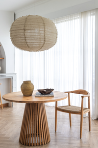 Round wooden table with a vase and bowl, accompanied by a chair in a bright room with large windows.