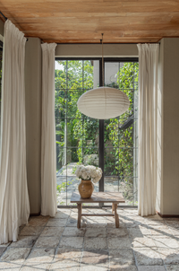 An oversized white dome shade hanging over a window, with a wooden chair and a vase of flowers in the foreground.