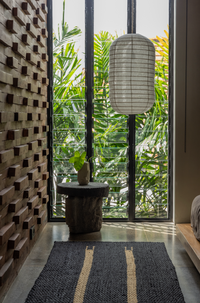 A narrow, capsule-shaped light shade hanging from a ceiling in a well-lit room with a lush green view. The shade appears to be made of a solid black material with a geometric pattern.