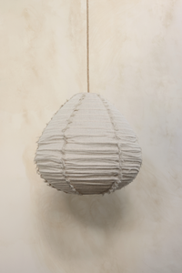 A pear-shaped linen light shade hanging against a wooden background, with a fringed design.